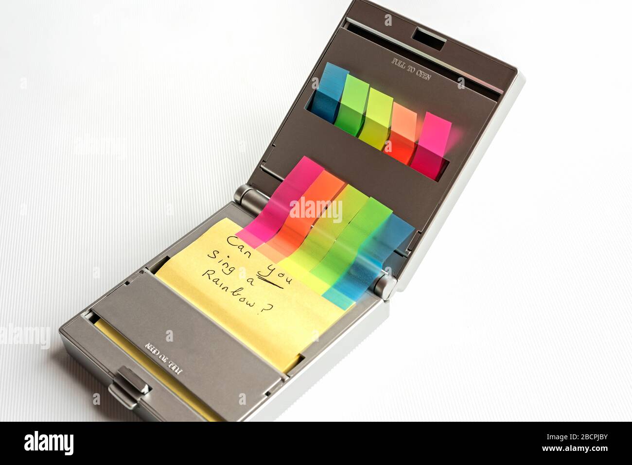 Silver dispenser of yellow postit notes & multi coloured sticky index highlighters & CAN YOU SING A RAINBOW? written on postit note, white background. Stock Photo
