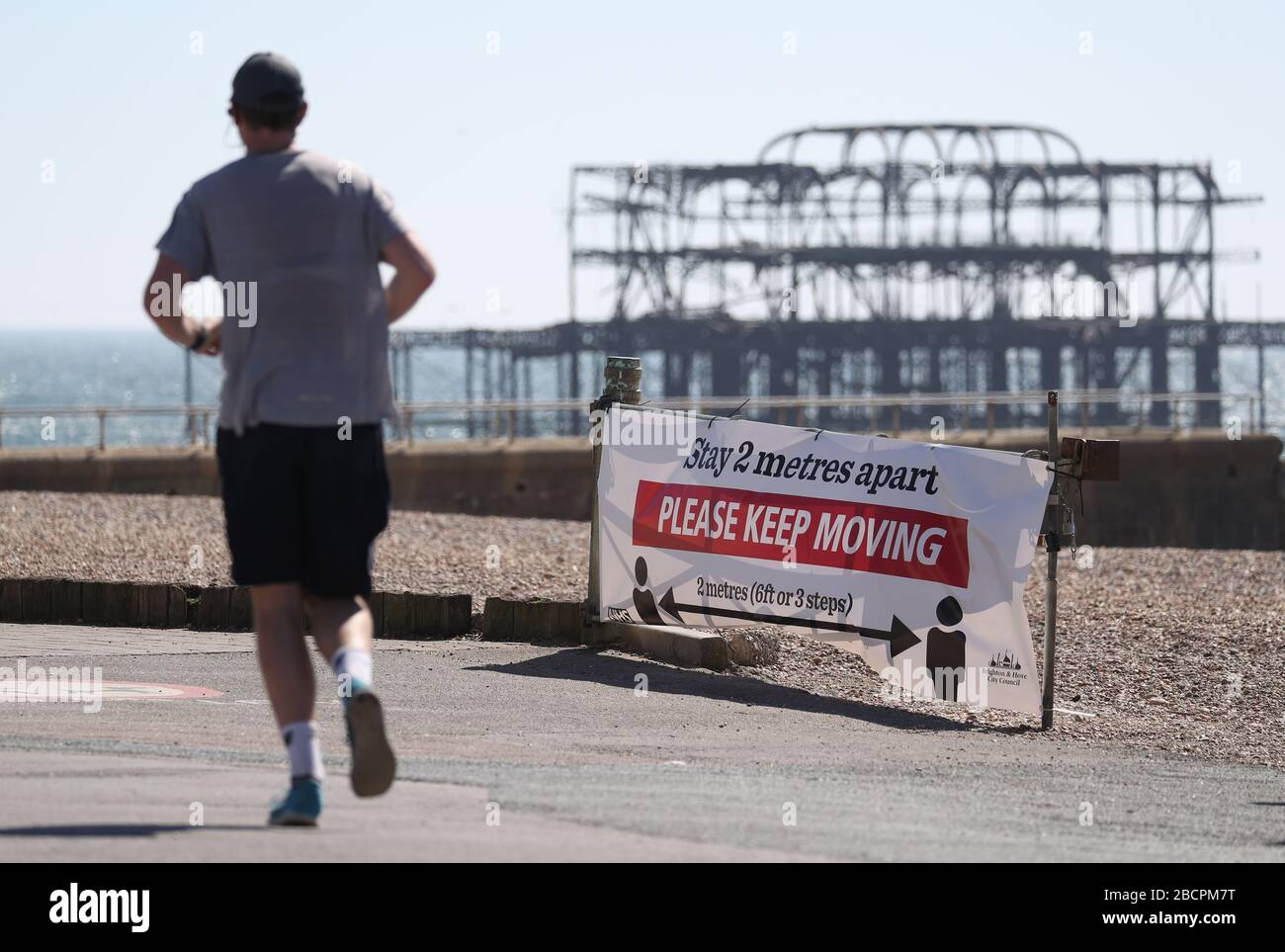 A jogger runs past a sign asking people to stay two metres apart on Brighton sea front, as the UK continues in lockdown to help curb the spread of the coronavirus. Stock Photo