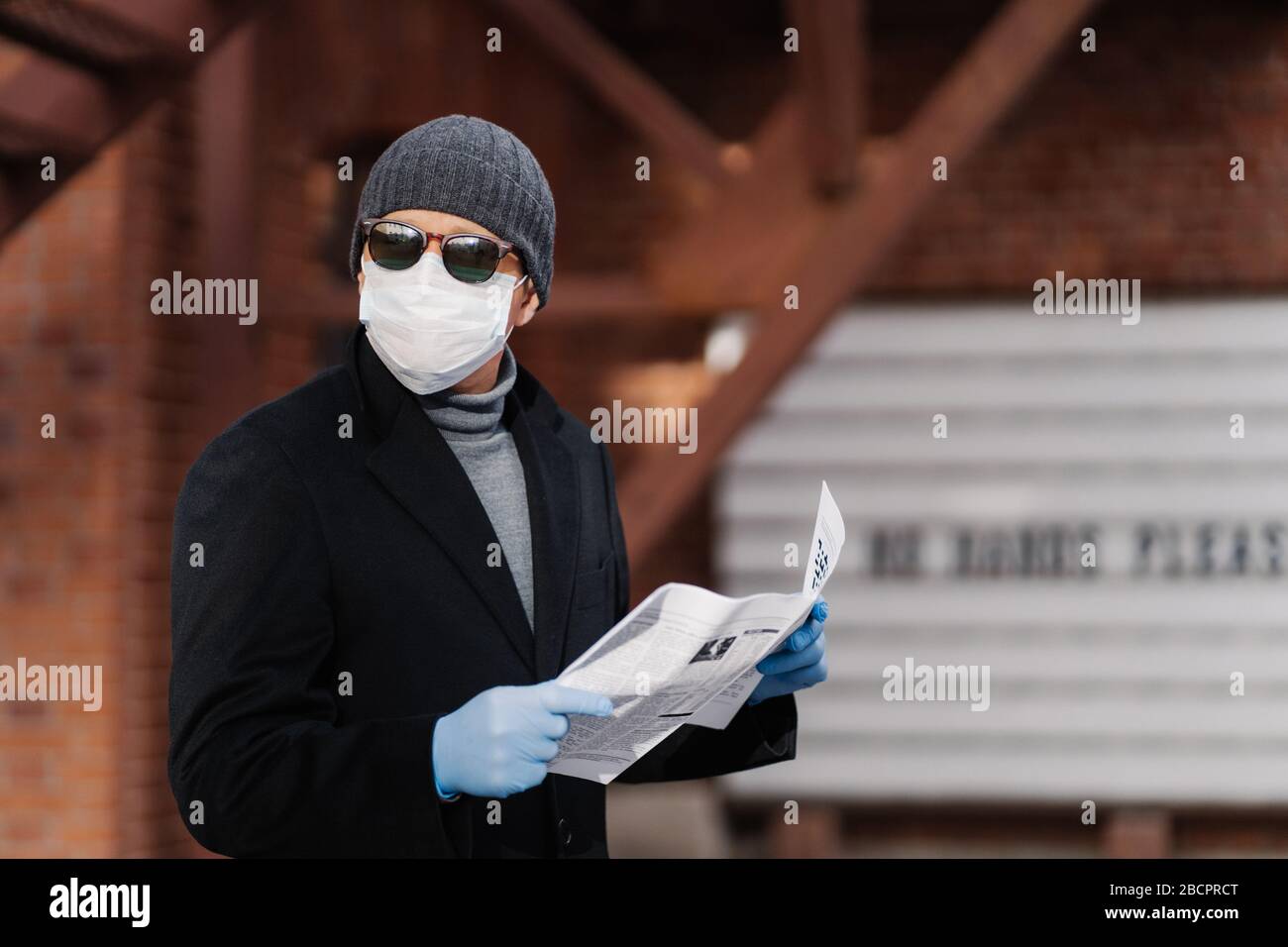 Horizontal shot of man uses virus preventive measures during coronavirus pandemic, wears face medical mask and rubber gloves, reads newspaper. Epidemi Stock Photo