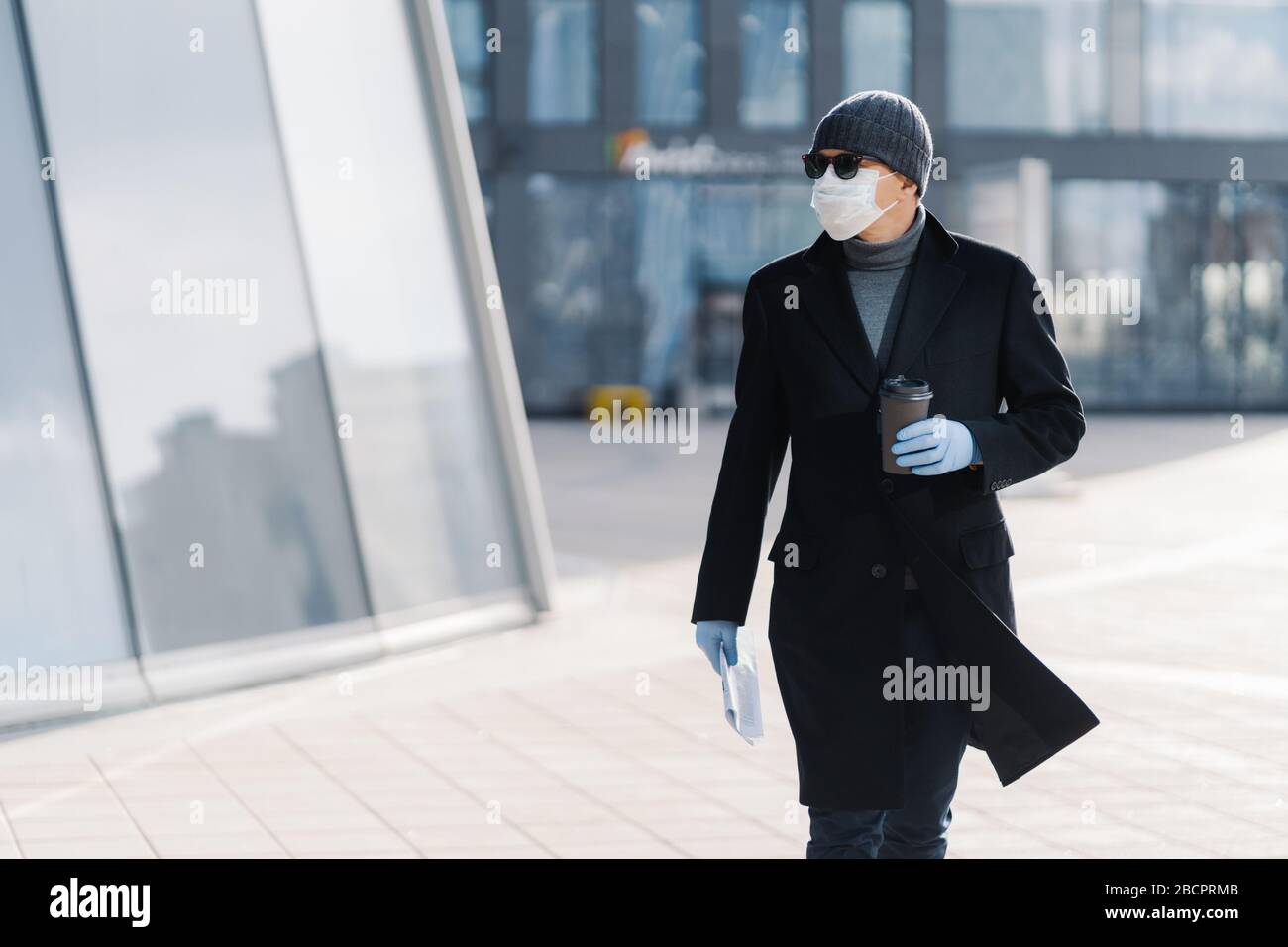 Horizontal shot of serious adult man carrying disposable cup of coffee, dressed in outerwear, protective medical mask and gloves, protects himself dur Stock Photo