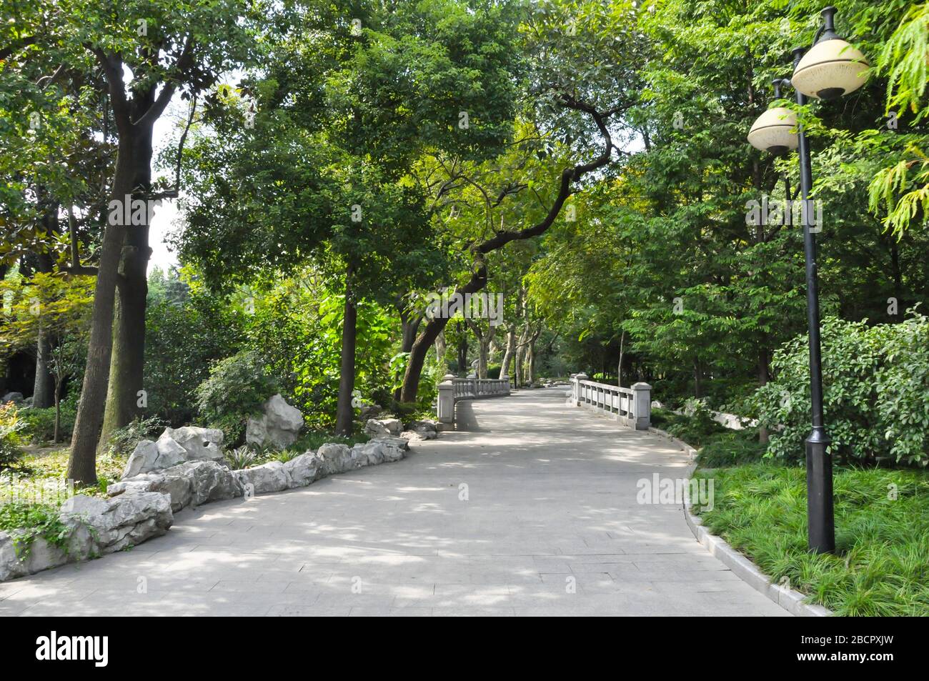 A concrete path through a park in the French concession in Shanghai, China. The path is surrounded by trees and lanterns and leads to a small white br Stock Photo