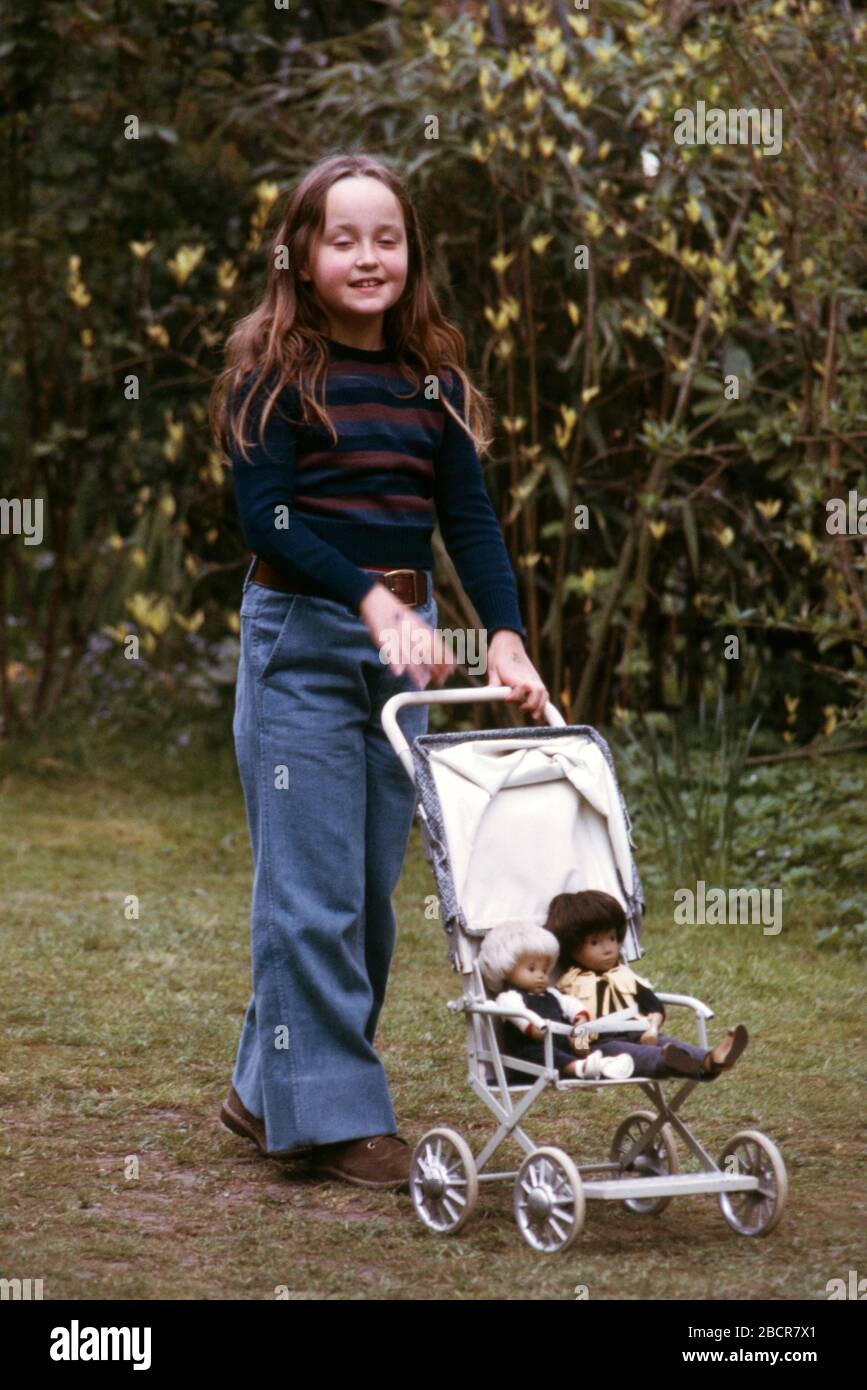 A 9 year old girl with long dark hair wearing blue denim jeans in garden of her family home playing with her Sasha dolls Gregor and Joe in a pushchair 1973 UK Stock Photo