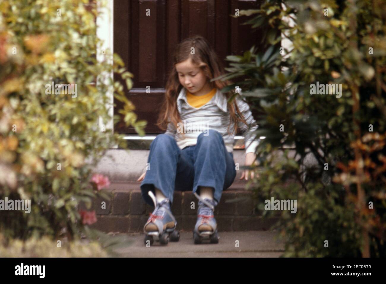 A 9 year old girl with long dark hair wearing blue denim jeans in the front garden of her family home, sitting on the step just about to get up onto her roller skates 1974 UK Stock Photo