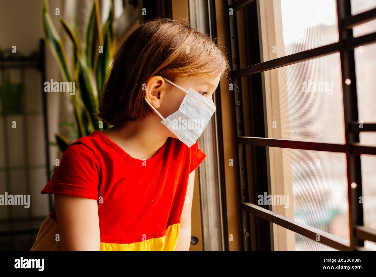 Small child with virus protection surgical face mask looking out the window, staying home for social distancing due to coronavirus Stock Photo