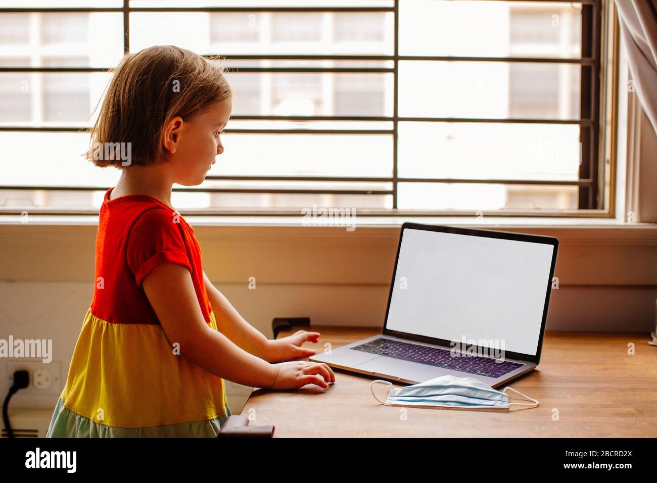 Portrait of a small preschool child with laptop computer and surgical face mask, using internet online at home by the window Stock Photo