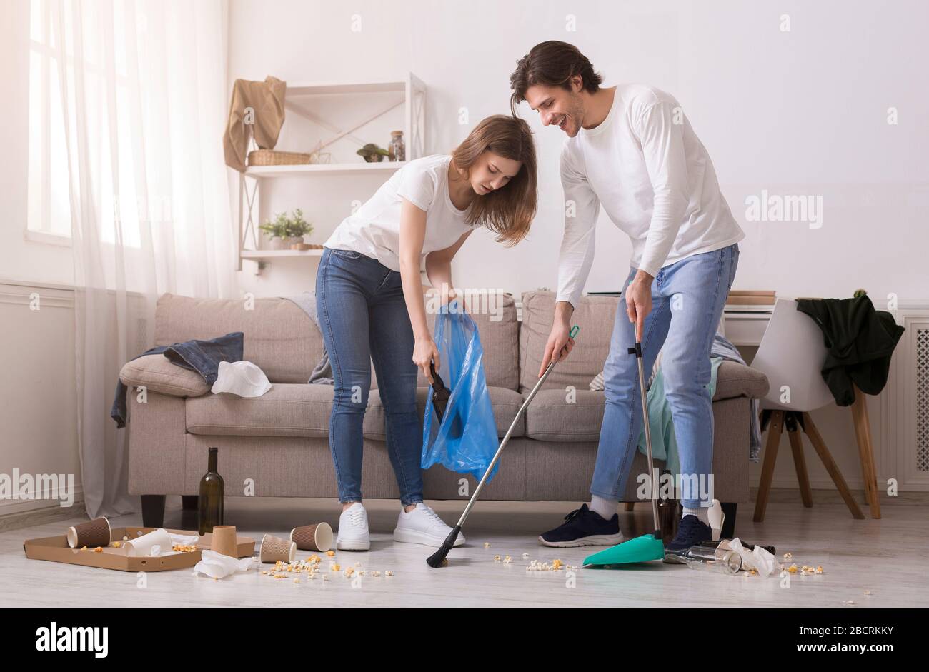 Couple Cleaning Messy Room After Party, Sweeping Floor And Collecting Garbage Stock Photo