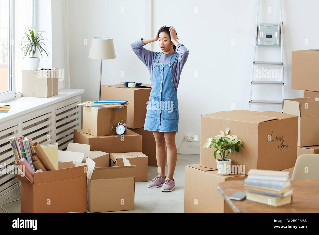 Full length portrait of frustrated Asian woman panicking while standing among cardboard boxes in empty room, house moving or relocation concept, copy Stock Photo
