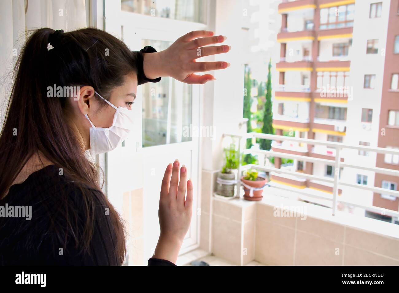 Asian woman doctor wearing a medical mask is looking out of the window with longing. The coronavirus spends quarantine days at home. Stock Photo