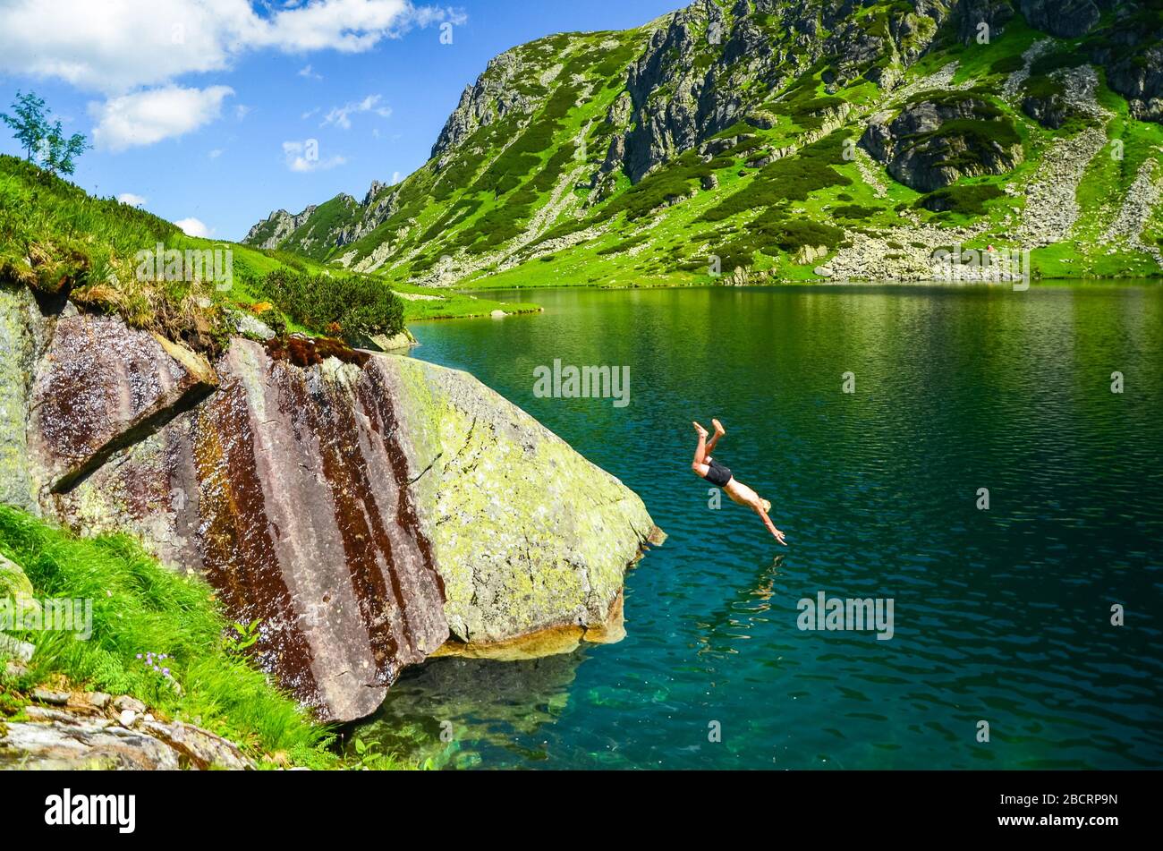 Two young man jumping to the beautiful blue lake in mountains. Summer scenery full of freedom in beautiful landscape Stock Photo