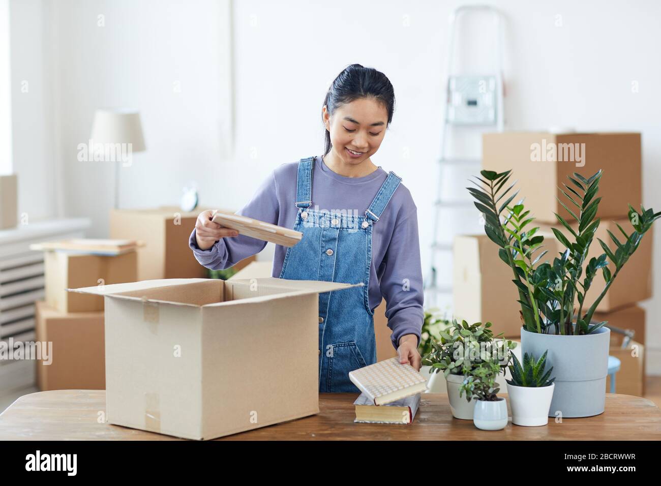 Waist up portrait of young Asian woman packing or unpacking cardboard box and smiling happily while moving into new home, copy space Stock Photo