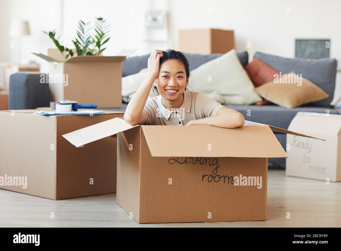 Portrait of young Asian woman sitting in box and smiling at camera while moving in to new house or apartment, copy space Stock Photo