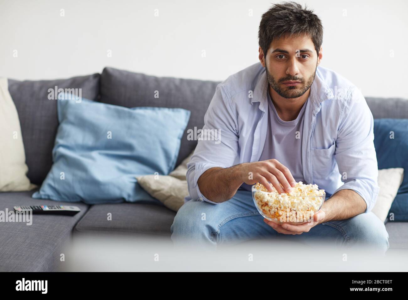 Portrait of sad bearded man watching TV and holding bowl of popcorn while sitting on sofa at home, copy space Stock Photo
