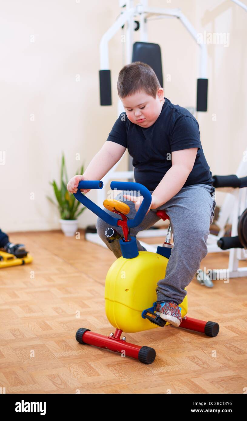 cute kid with down syndrome exercising on stationary bike at the inclusive sport center Stock Photo