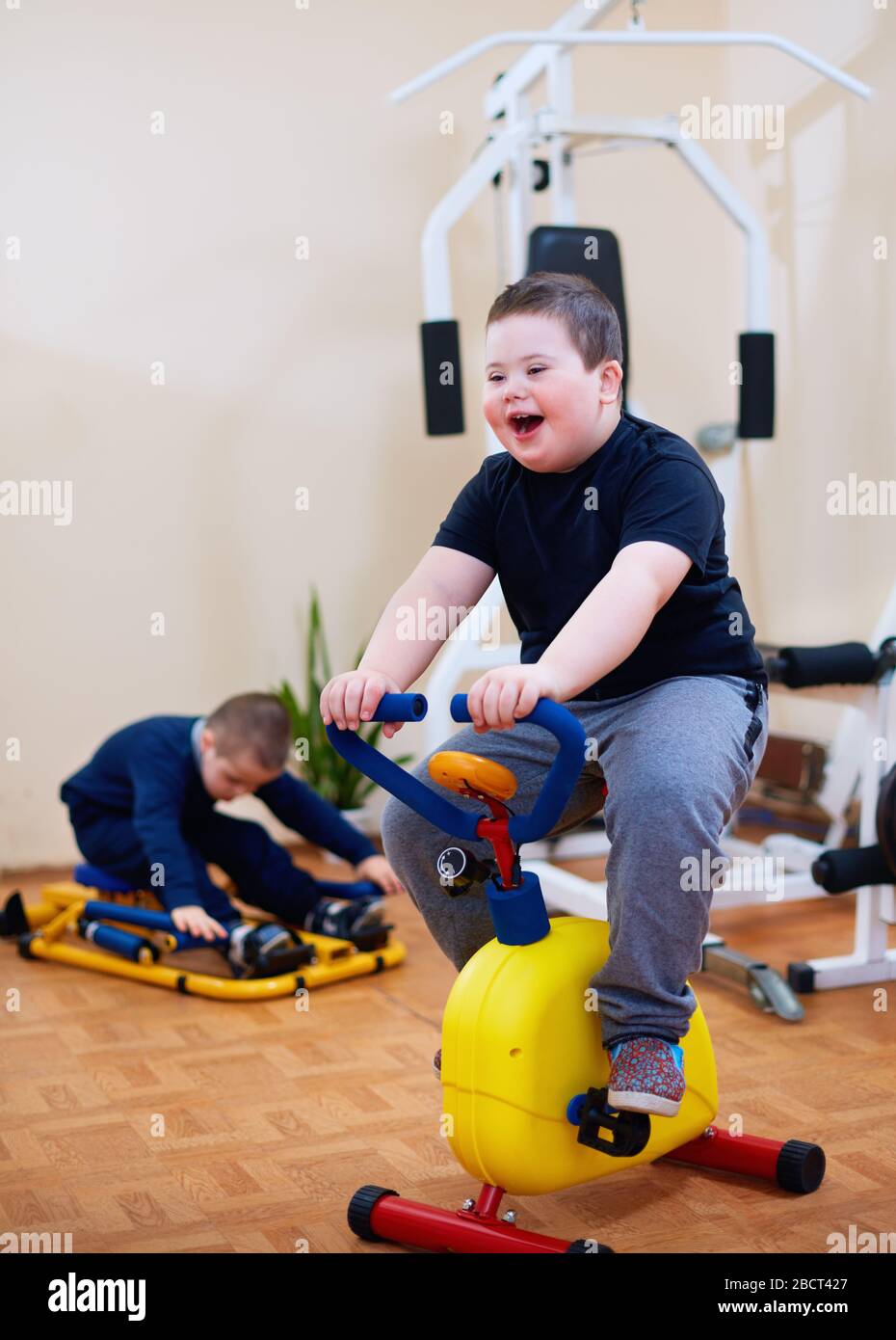 cute cheerful kid with down syndrome exercising on stationary bike at the inclusive sport center Stock Photo