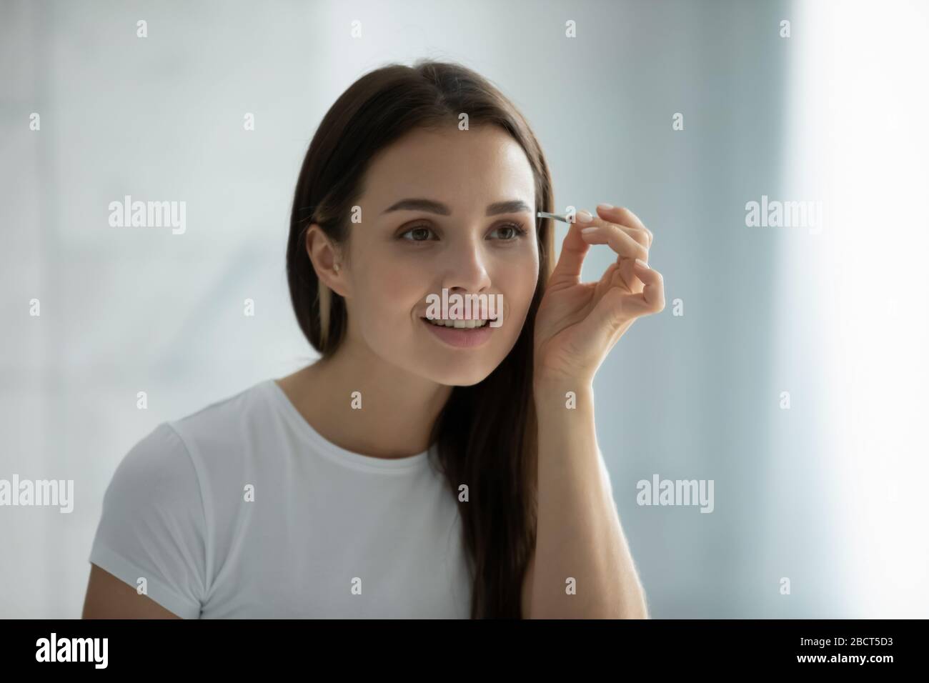 Young woman look in mirror pinching eyebrows with tweezers Stock Photo