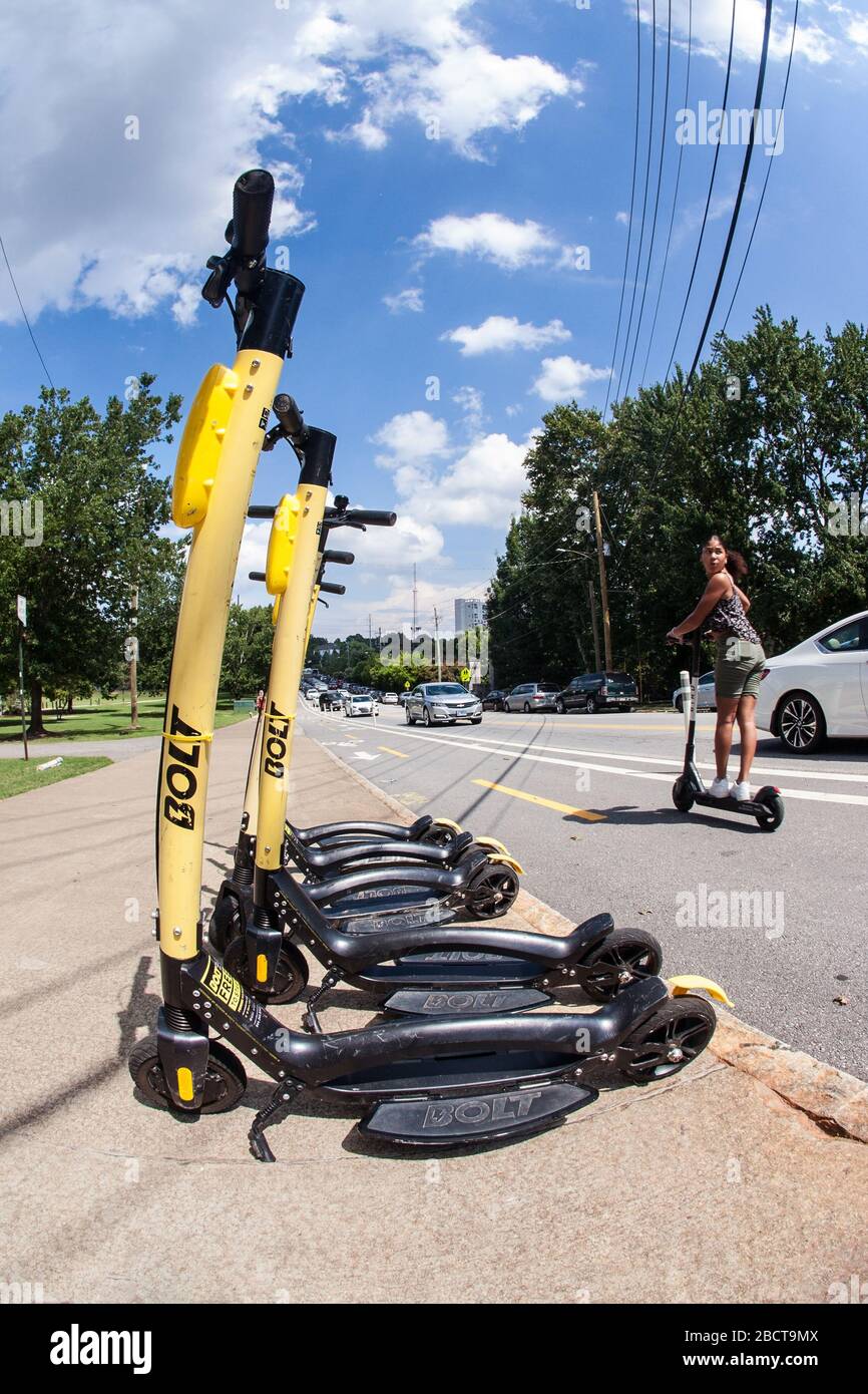 A young woman riding a motorized scooter passes by several parked scooters on the sidewalk at Piedmont Park on August 10, 2019 in Atlanta, GA. Stock Photo