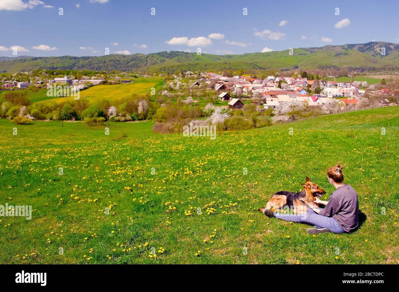 Girl and dog are sitting on spring meadow and they are looking to wonderfull village which is full of colors and pleasure afternoon light. They love e Stock Photo