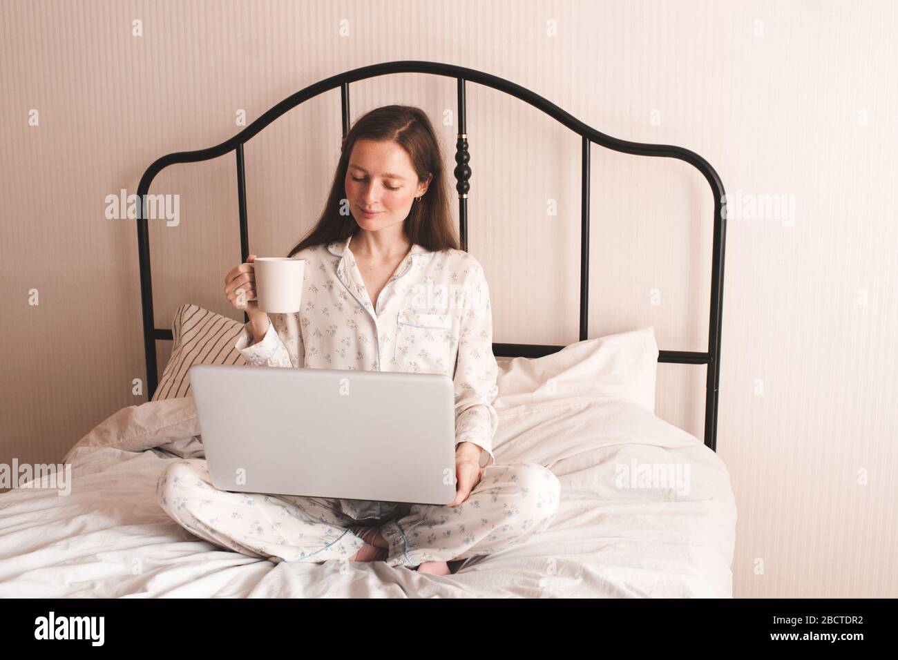 Young woman drinking fresh tea and holding laptop sitting in bed and working at home indoors. Good morning. Breakfast time. Freelance. Social distanci Stock Photo