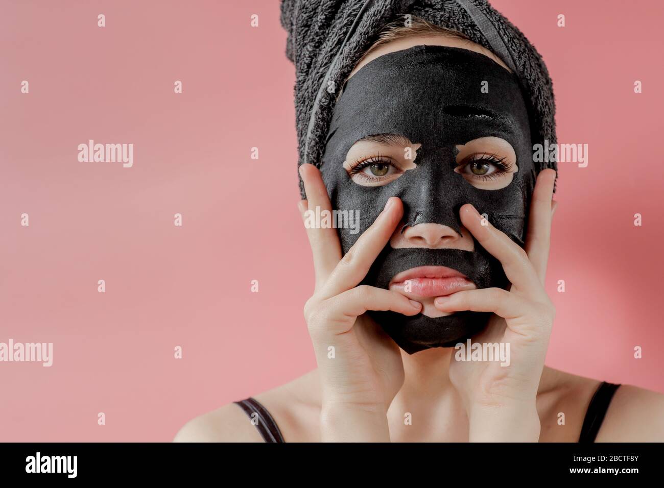 Young woman apply black cosmetic fabric facial mask on pink background