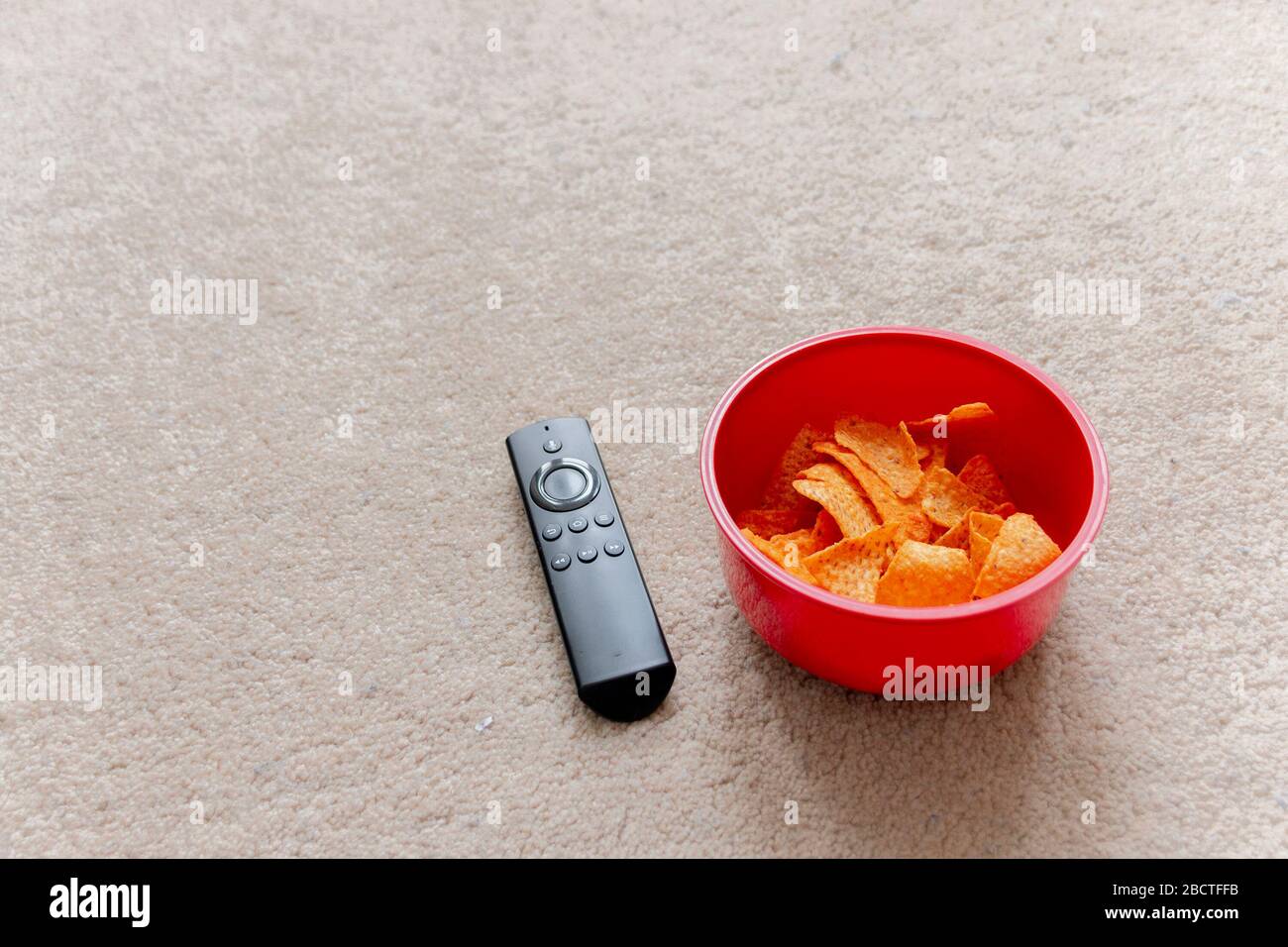 a close up view of a smart tv remote next to a red plastic bowl of chips or crisps on the light coloured carpet Stock Photo