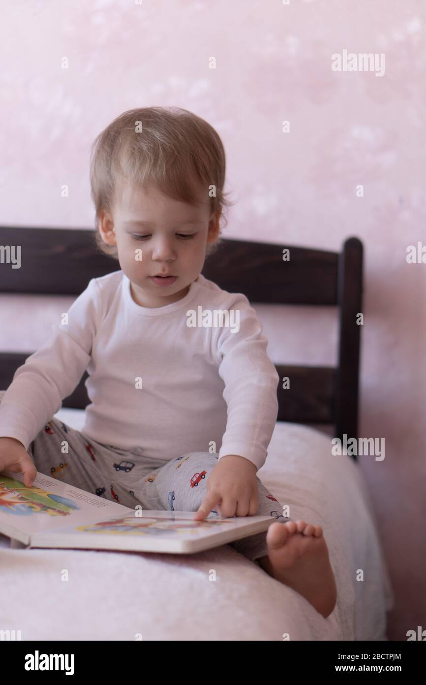 A little blond-haired boy is sitting on the bed and smiling, holding a book in his hands. A little boy sits on a bed with a book in his hands during Stock Photo