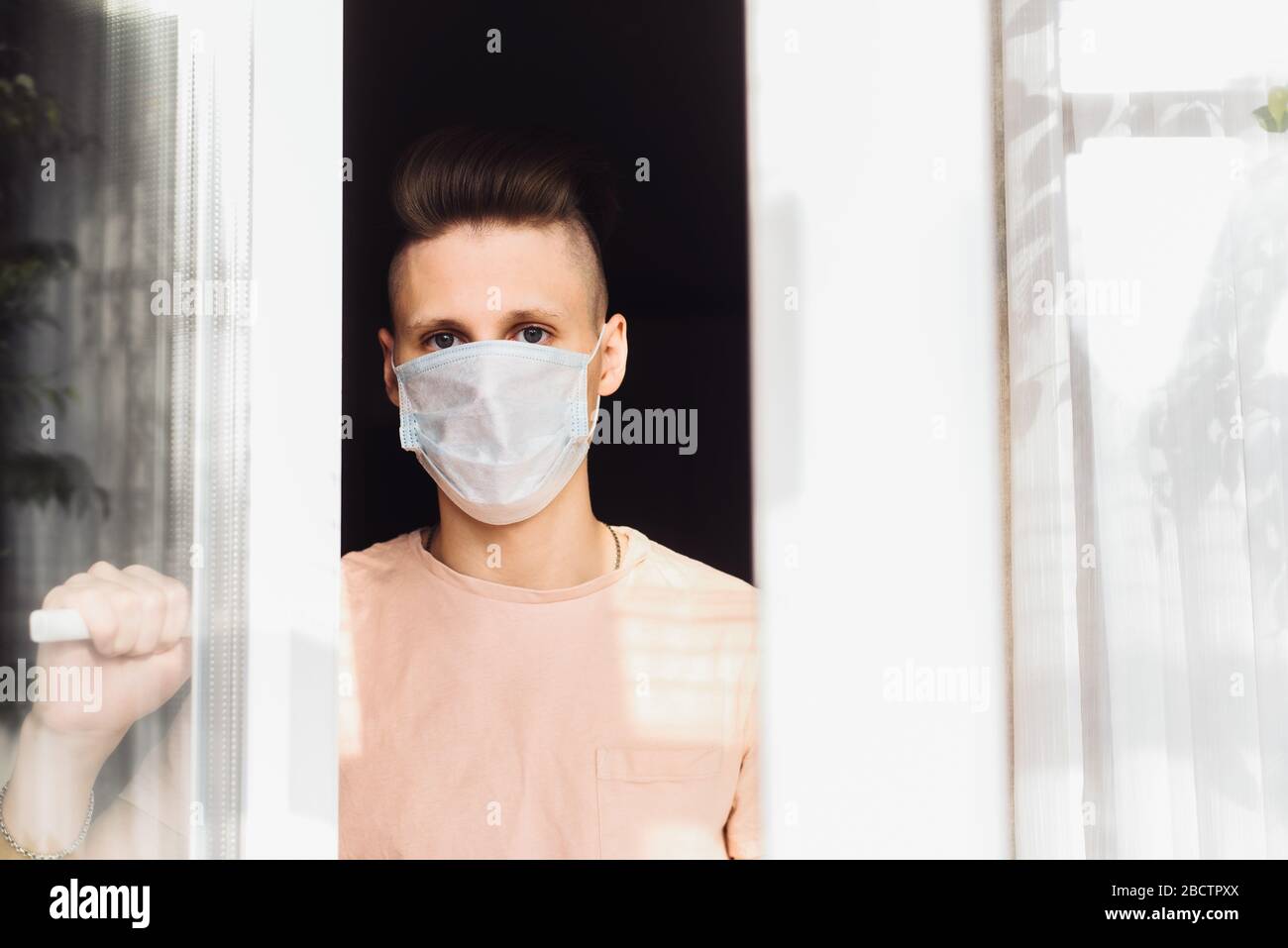 Handsome young man in a protective mask stands at home outside the window. A call for self-isolation due to a pandemic and the risk of coronavirus infection. Stock Photo