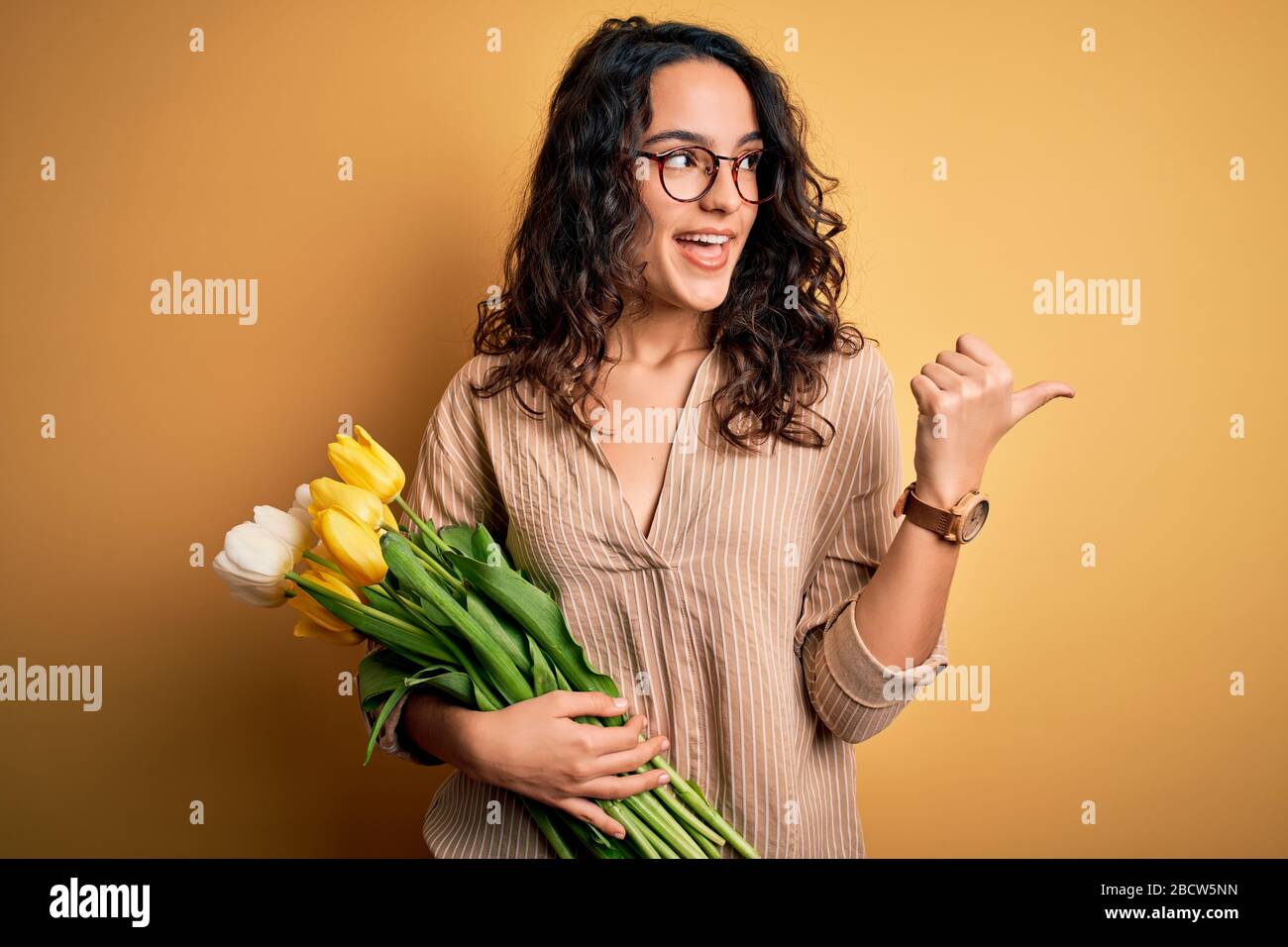 Young beautiful romantic woman with curly hair holding bouquet of yellow tulips smiling with happy face looking and pointing to the side with thumb up Stock Photo