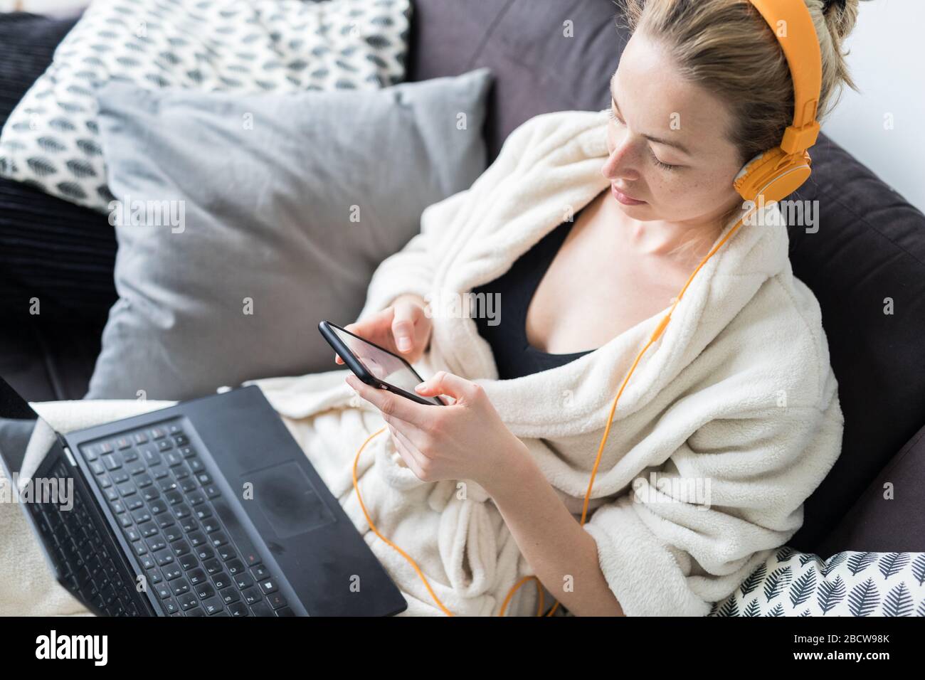 Social distancing. Stay at home. Woman in bathrobe being comfortable at her home sofa, using social media apps on phone for video chatting and stying Stock Photo