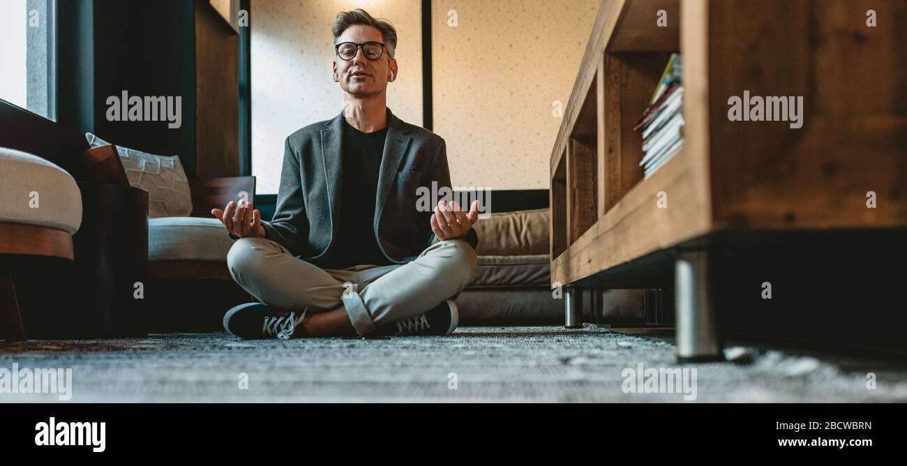 Mature businessman doing yoga meditation in office lounge. Male business executive meditating in lotus pose on the floor in the office. Stock Photo