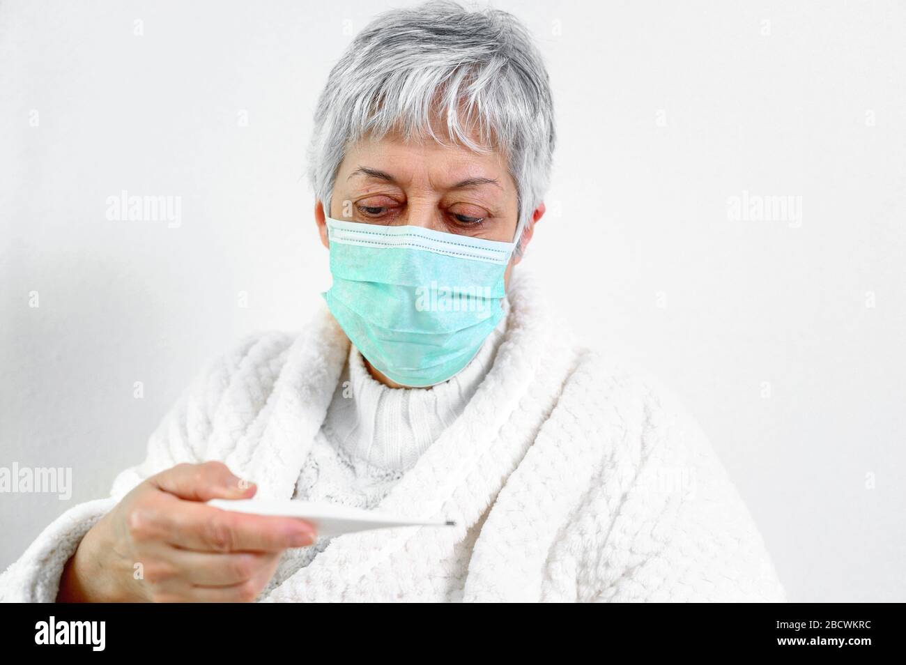 Coronavirus. Sick senior woman holding thermometer sit on bed, upset old mature woman caught cold got coronavirus flu influenza symptoms at home alone . Stock Photo