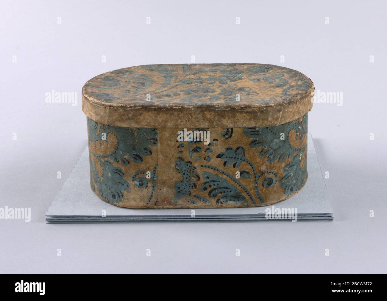 Bandbox. Research in Progressa) Oval box covered with wallpaper. Foliate scrolls in lavender-blue with darker blue dots emphasizing the pattern in various places, on yellow ground. Margin visible at seam; b) Cover of the same as 'a'. Two joined sheets. Bandbox Stock Photo