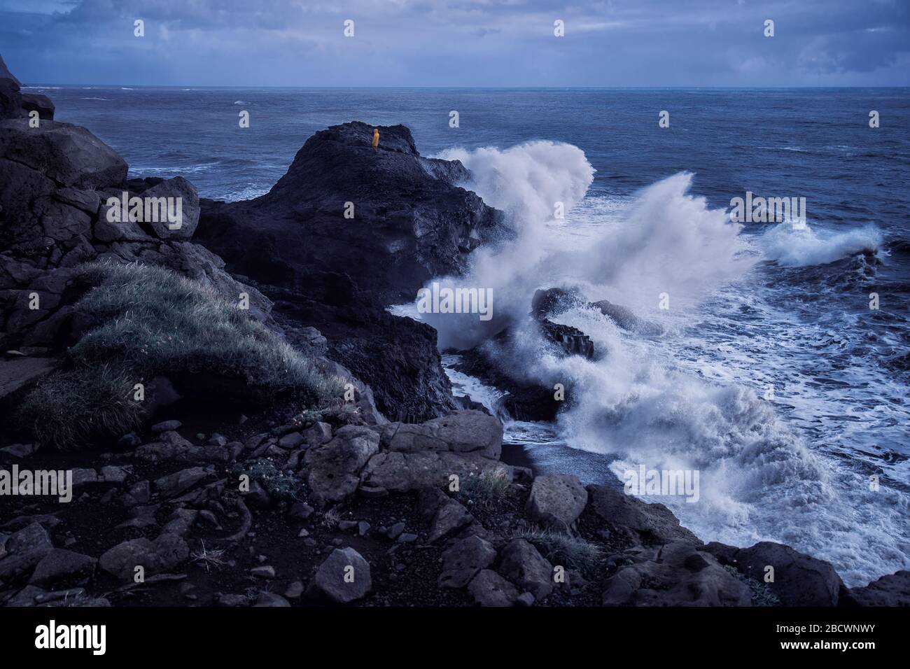 man in yellow raincoat standing at edge of cliff with huge waves breaking against it on stormy day in Dyrholaey, Iceland Stock Photo