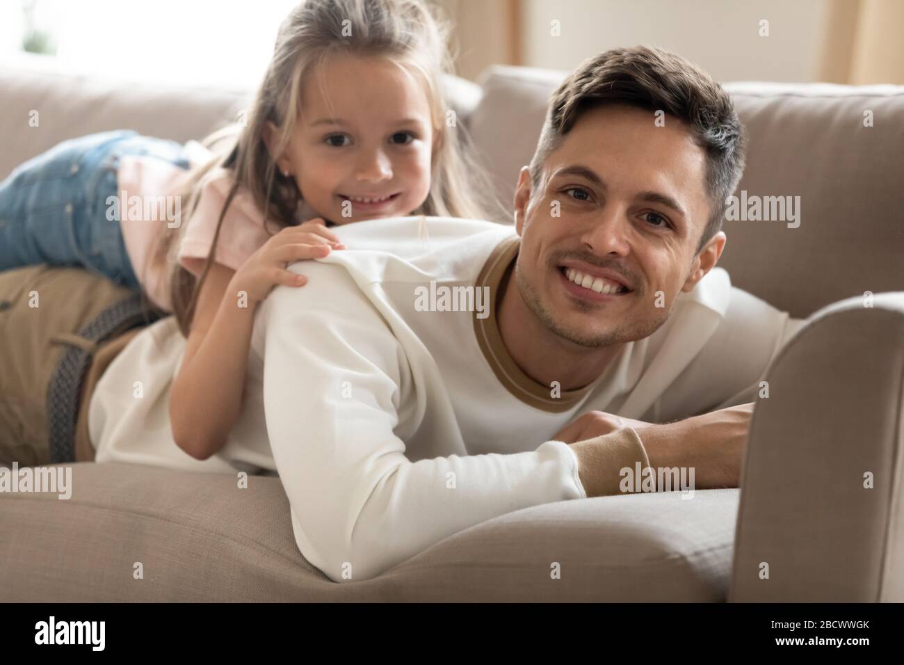 Happy young father lying on couch with smiling small daughter. Stock Photo