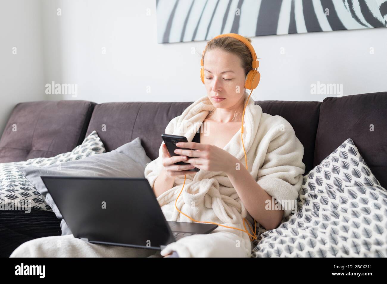 Social distancing. Stay at home. Woman in bathrobe being comfortable at her home sofa, using social media apps on phone for video chatting and stying Stock Photo