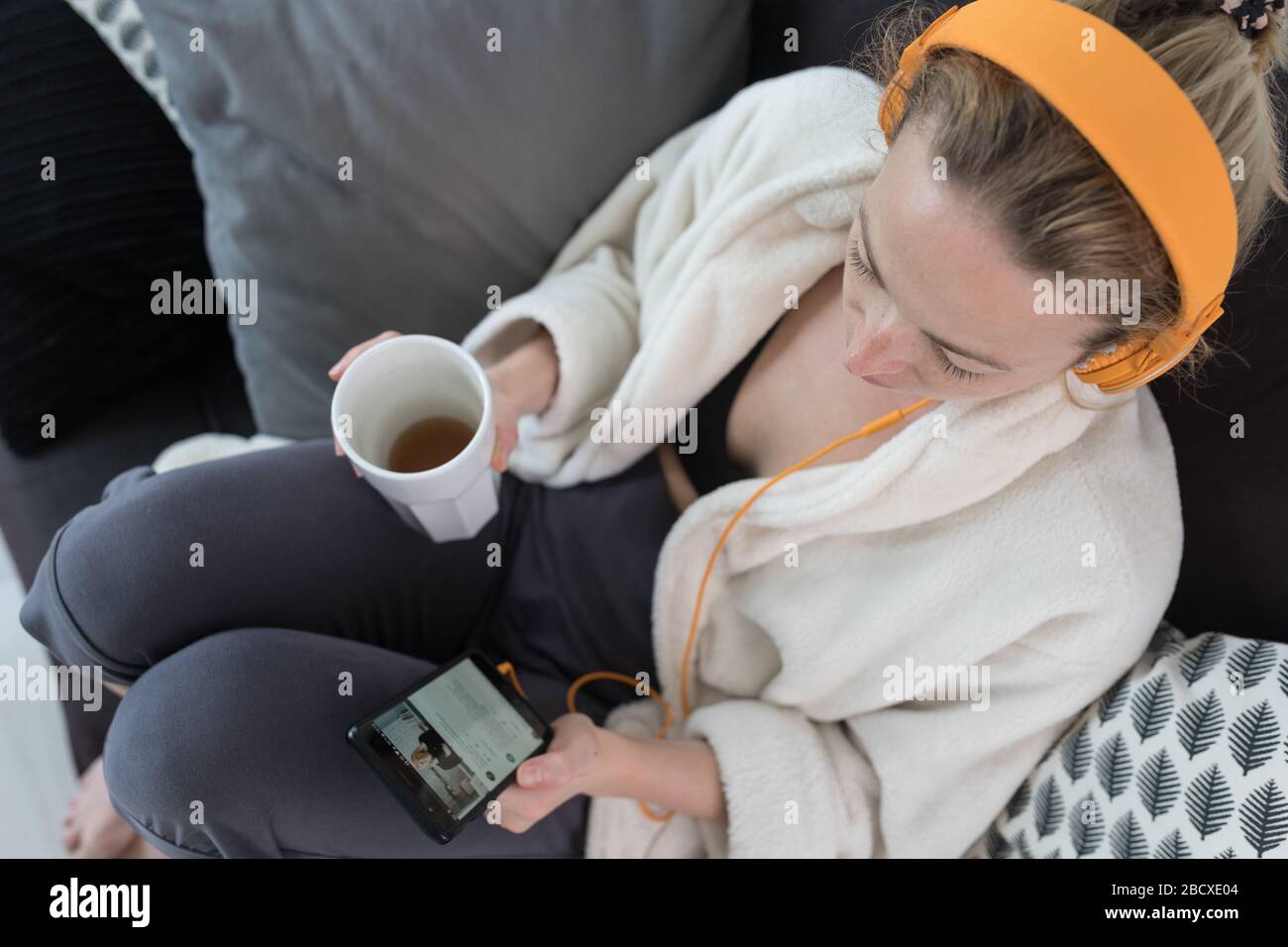 Stay at home. Social distancing. Woman at home relaxing on sofa couch drinking tea from white cup, listening to relaxing music, stay connected to Stock Photo