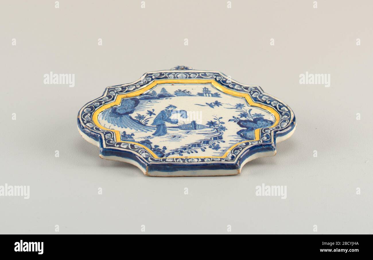 Plaque. Research in ProgressShaped plaque with raised molded border and circular ribbed hanging loop on top edge; painted in underglaze blue and yellow with leaf scroll border, chinoiserie scene center with figure in landscape. Plaque Stock Photo