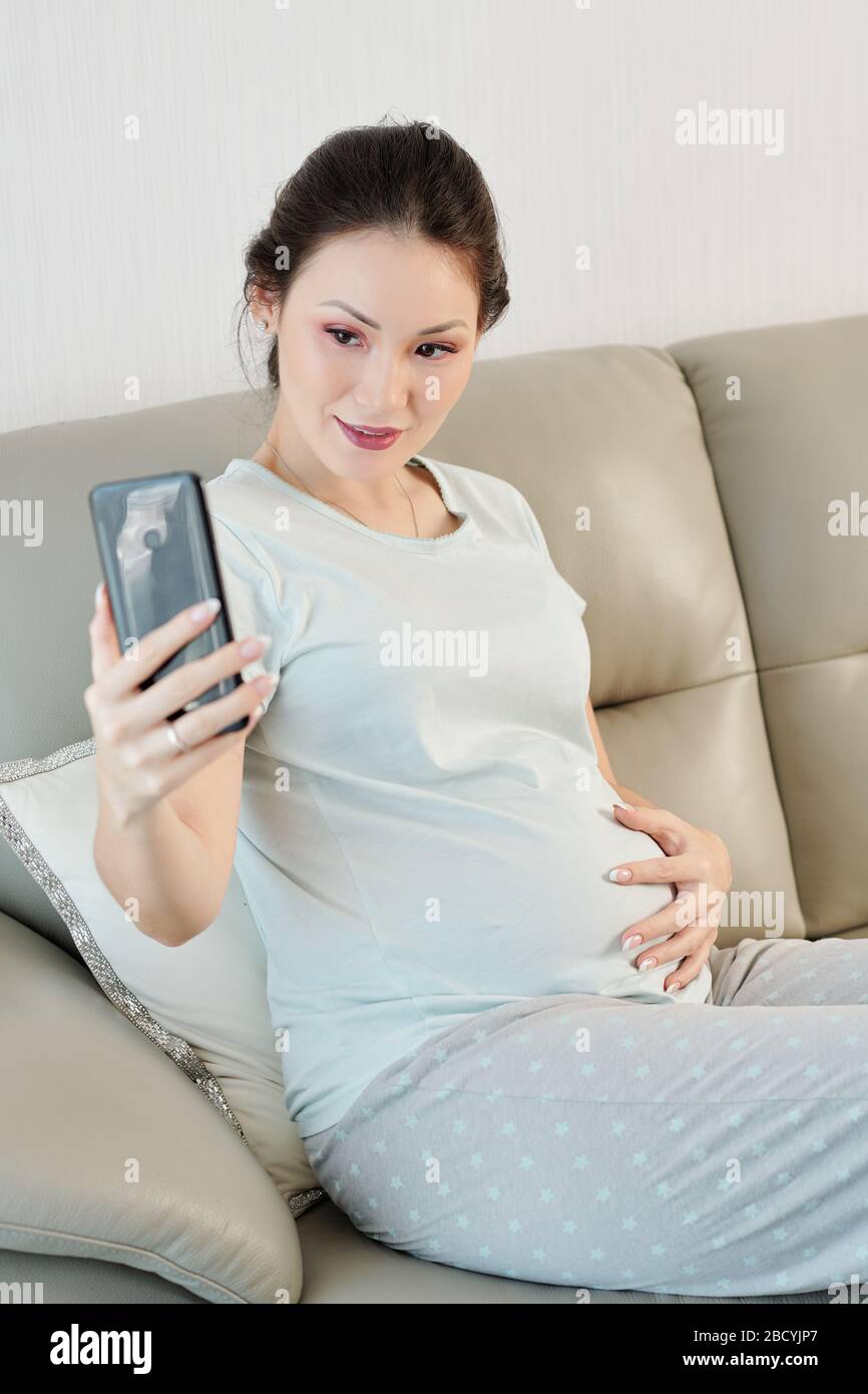 Beautiful young Vietnamese pregnant woman sitting on sofa and taking