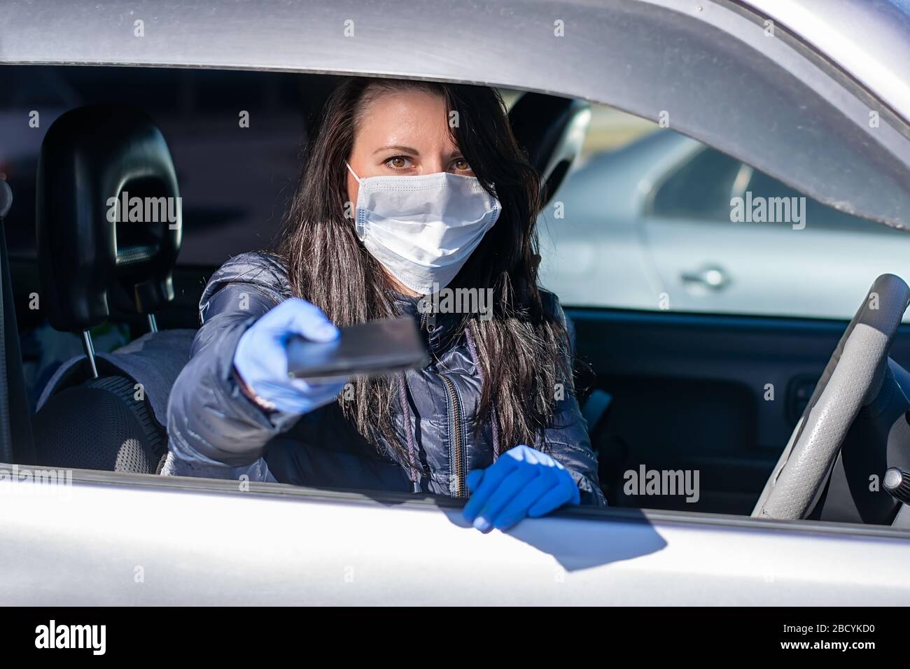 the girl driver, sitting in the car in a medical anti virus anti bacterial mask and rubber gloves, holds out documents for verification Stock Photo