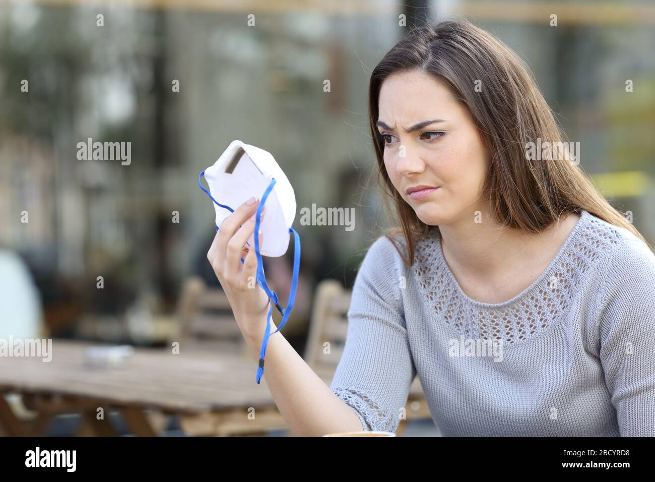 Doubtful woman looking hesitant at protective mask sitting on a coffee shop terrace Stock Photo