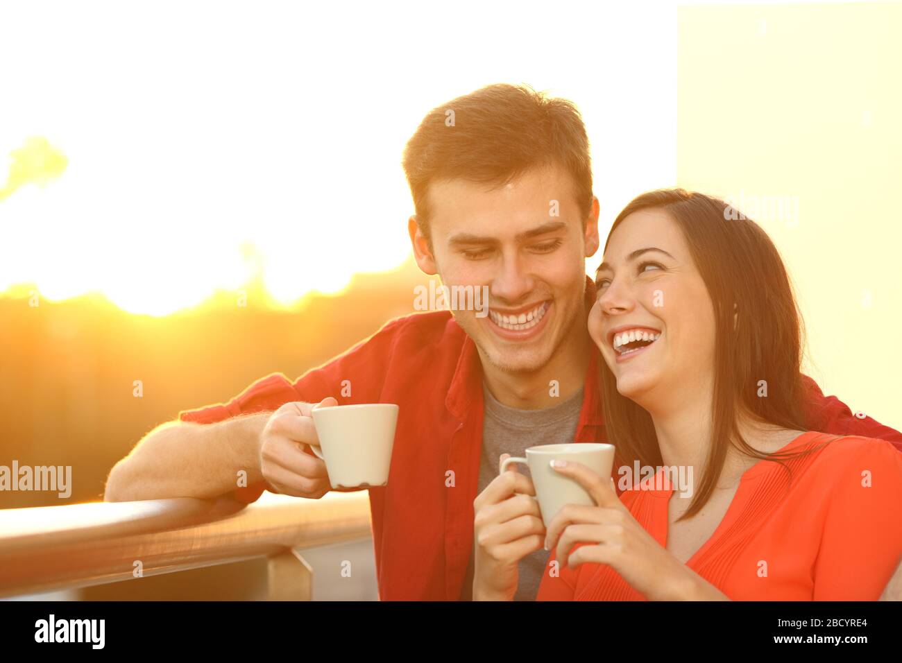 Happy couple cuddling holding coffee cups enjoying at sunset standing on a balcony at home Stock Photo