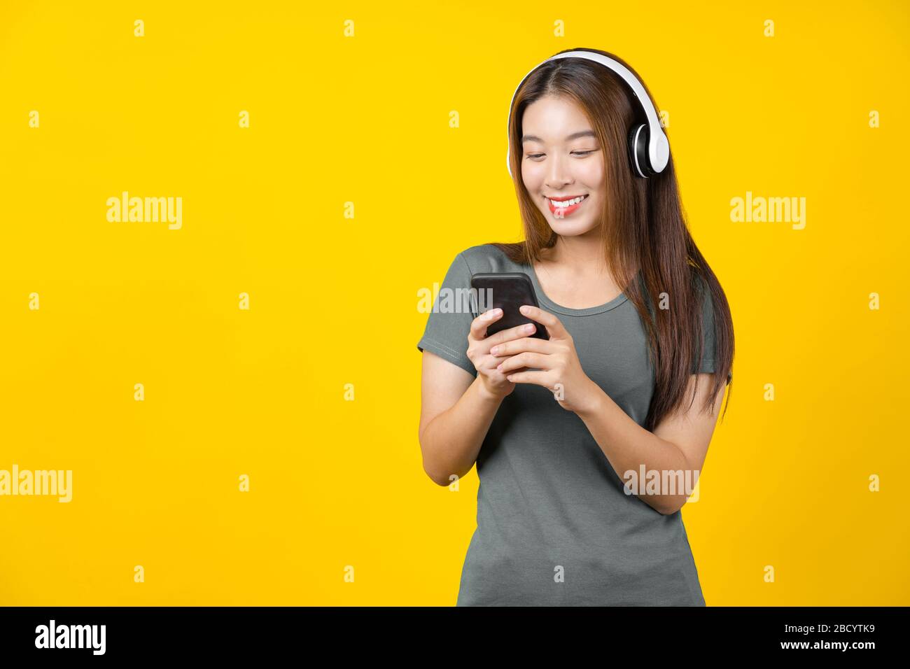 Happiness Asian smiling young woman wearing technology wireless headphones for listening the music via smart mobile phone on isolated yellow color bac Stock Photo