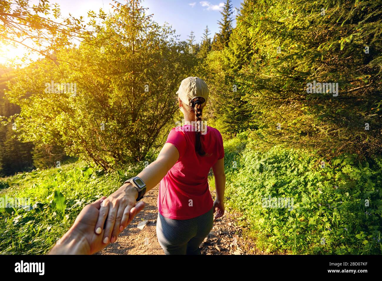 Beautiful sporty girl in pink shirt holding man by hand and leading him to the mountain trail at sunset Stock Photo