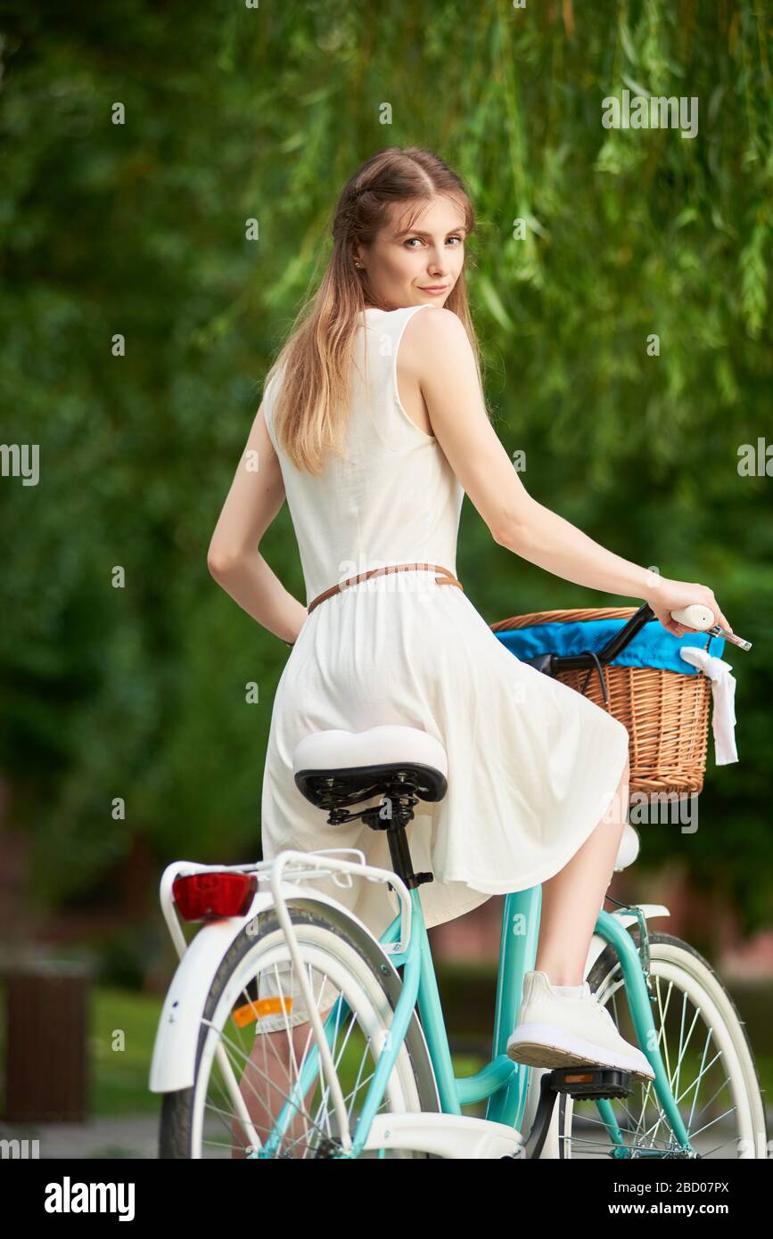Pretty woman on the retro bike with basket at blurred background greenery of city park. Girl is dressed in light dress and white sneakers posing with her back and turned to look at the camera. Stock Photo