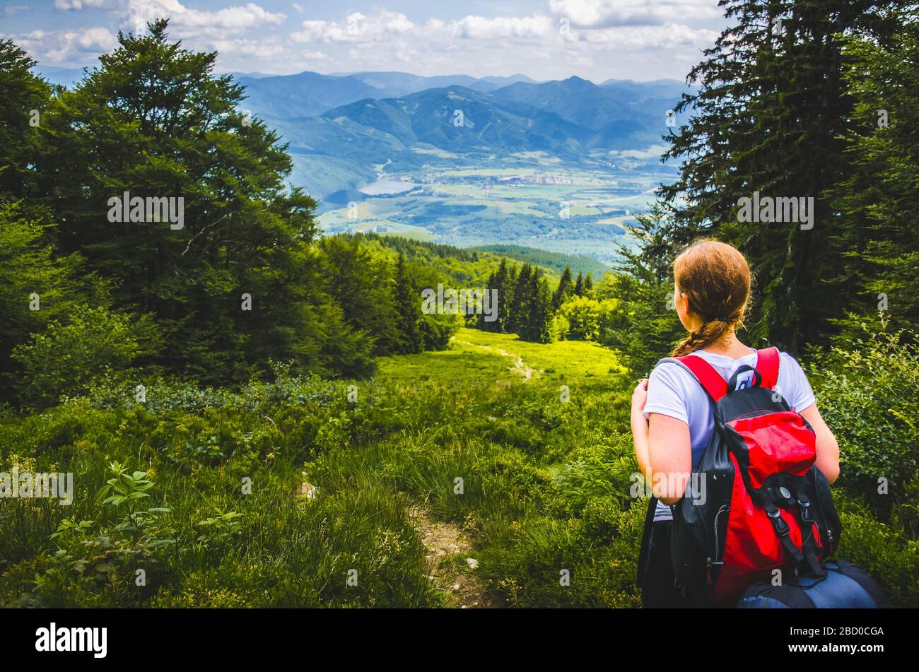 Alone female tourist with red rucksack walking up hill in green mountains under blue sky Stock Photo