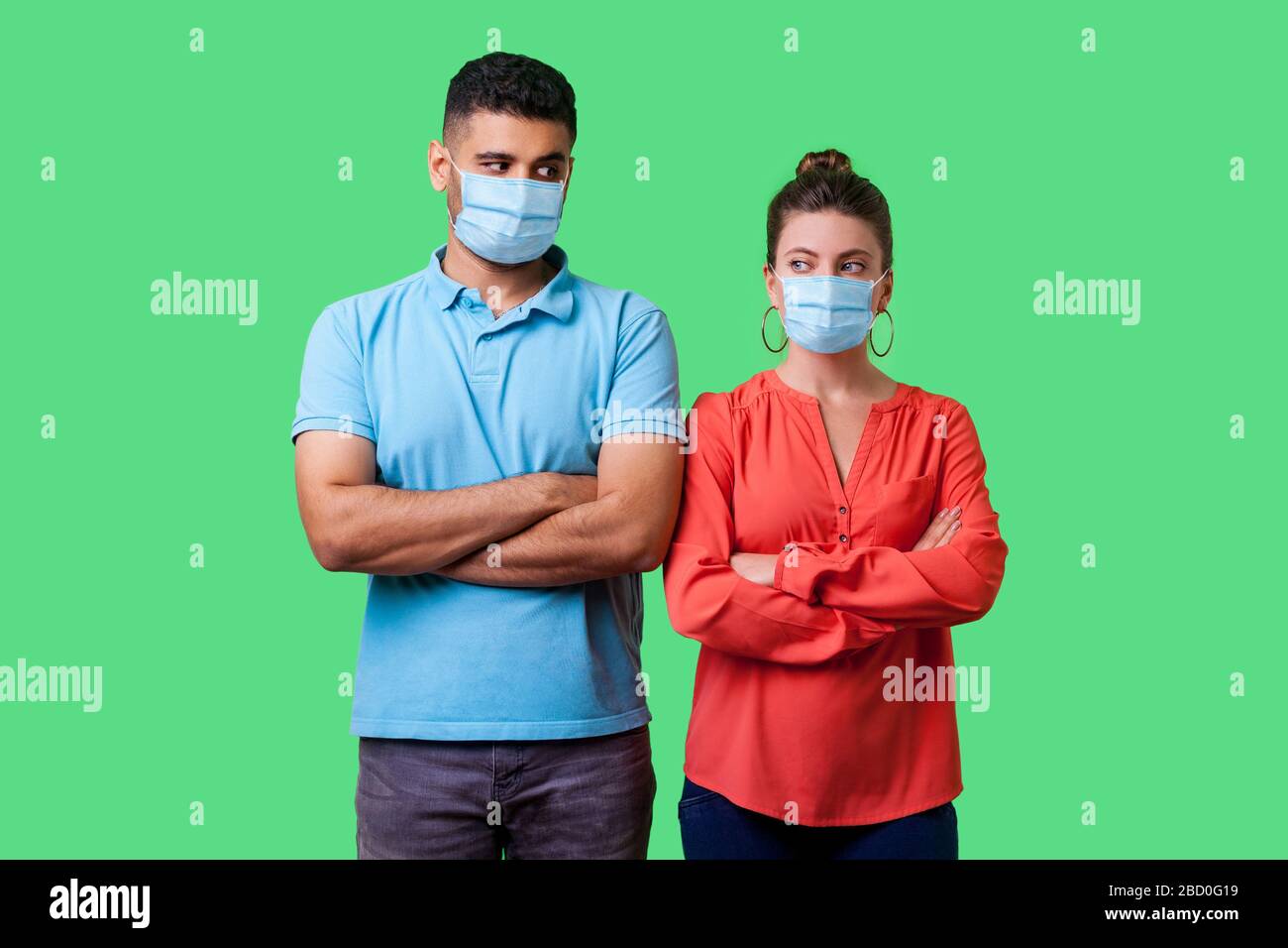 Portrait of upset couple with surgical medical mask standing together with crossed hands, looking sideways at each other with resentful glance, suspic Stock Photo