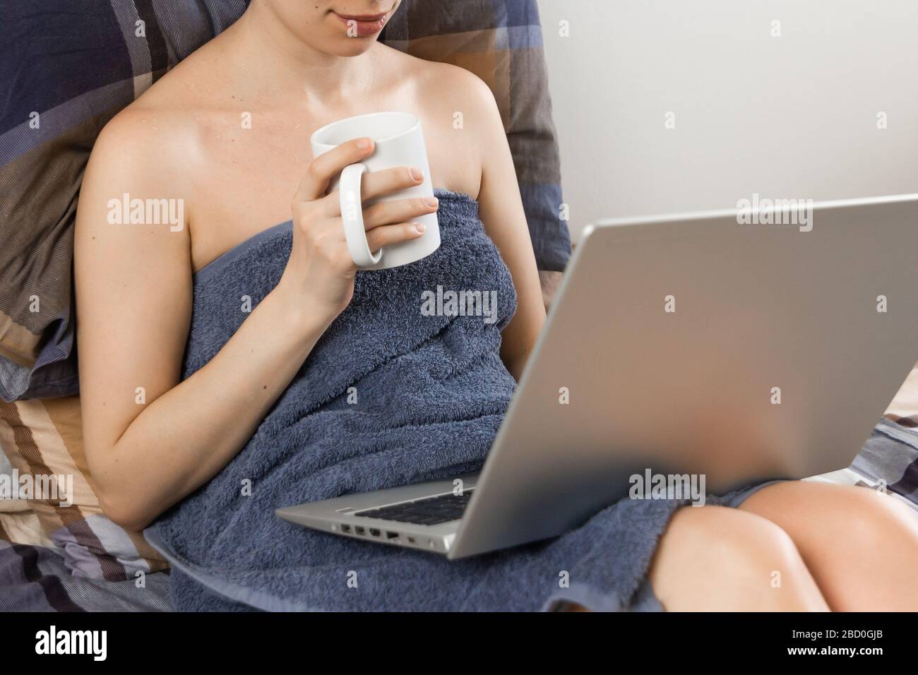 Freelancer woman is working on her laptop with cup of tea in the bed with blue linens. Concept of home office. Woman relaxes after bath covered in Stock Photo