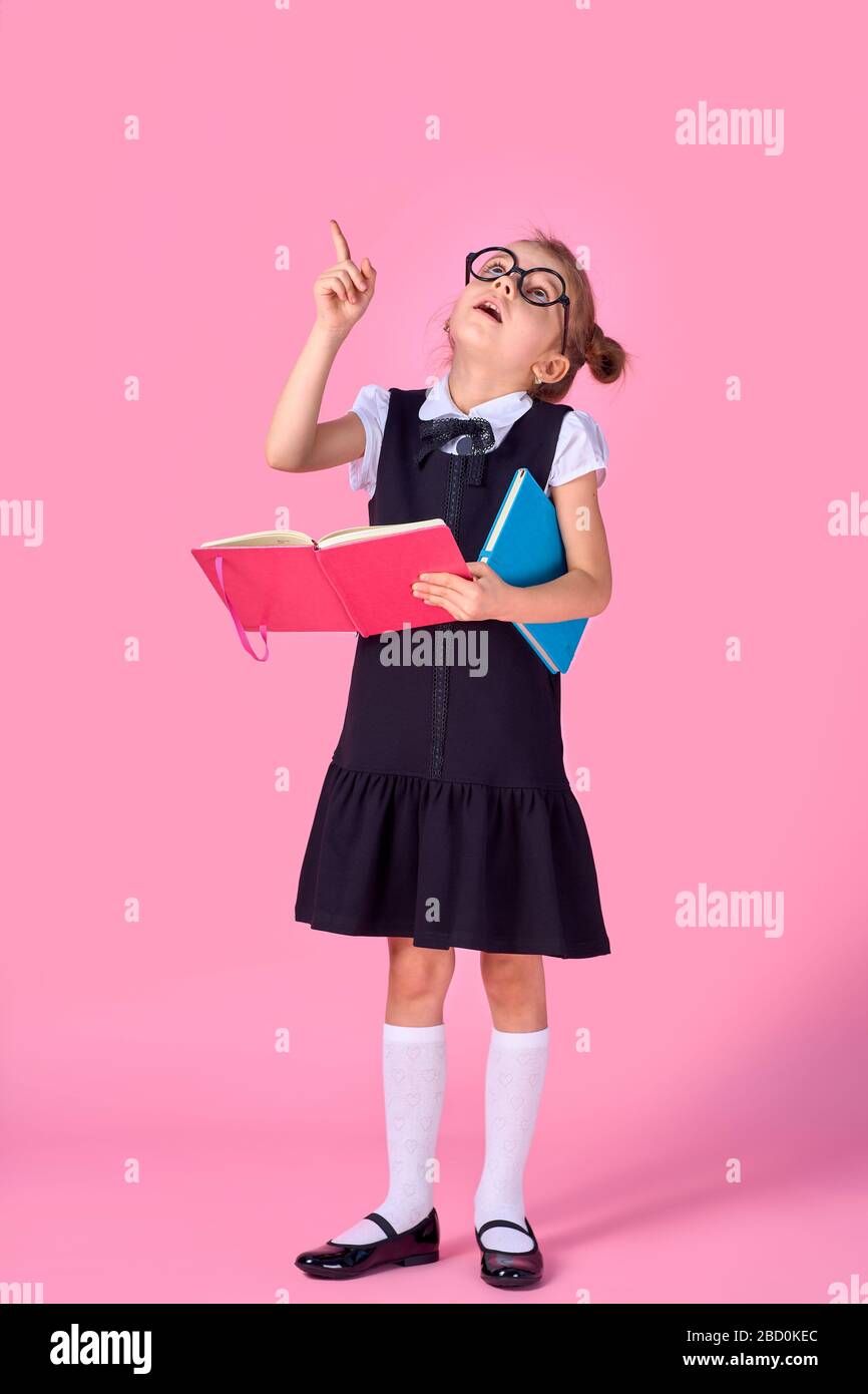 preschool girl with glasses holding a book, raised her hand and finger up on a pink background. Stock Photo