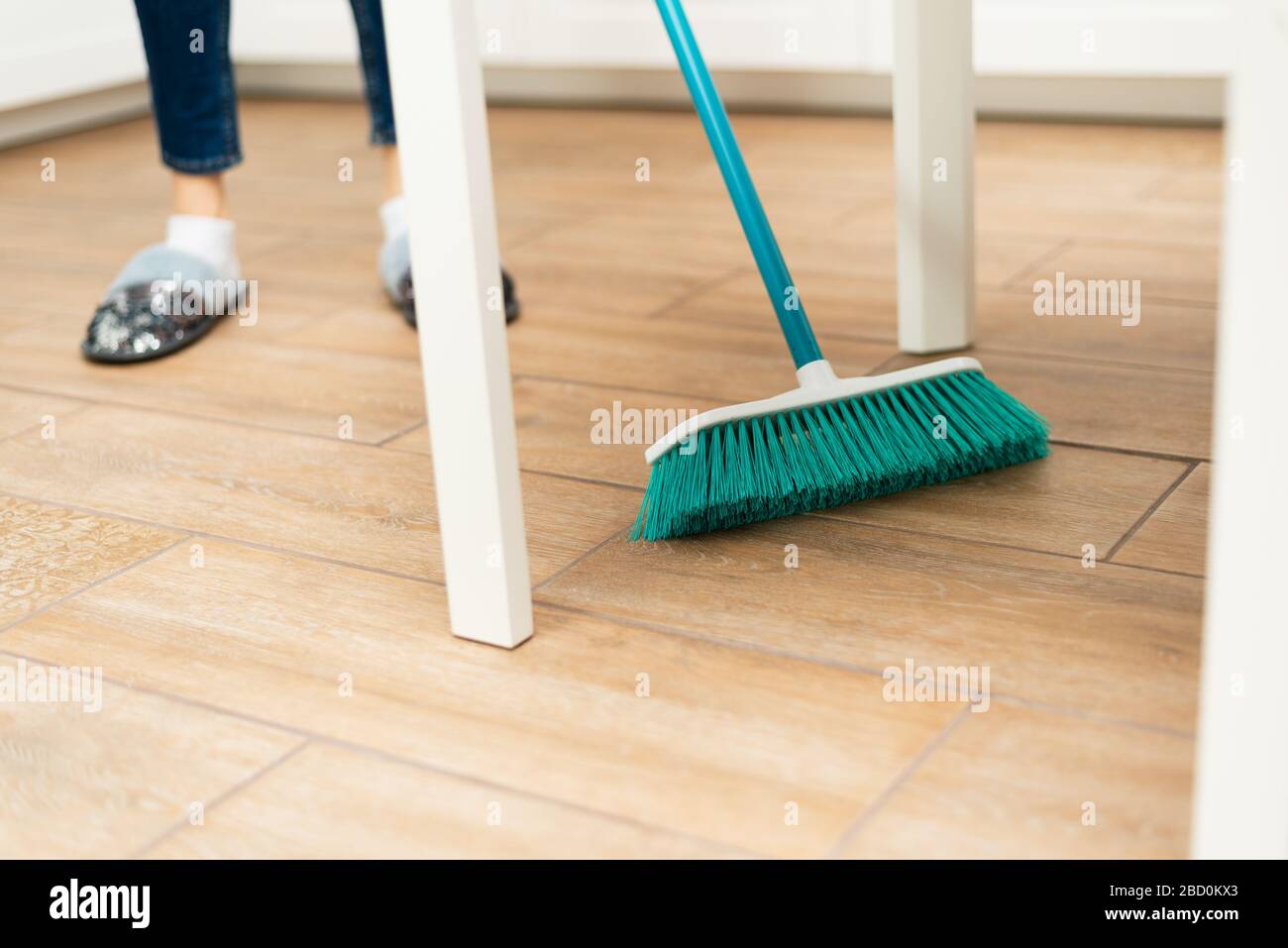 Young woman sweeps laminate flooring in a bright kitchen. The girl sweeps dust and dirt from under the white kitchen table. Stock Photo