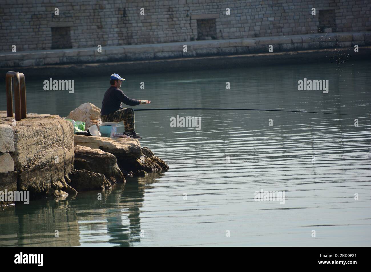 Trani, Italy-April 2019: lone man seated on some rocks doing some fishing in the harbor of Trani; fortification walls as background, swarm of mosquito Stock Photo