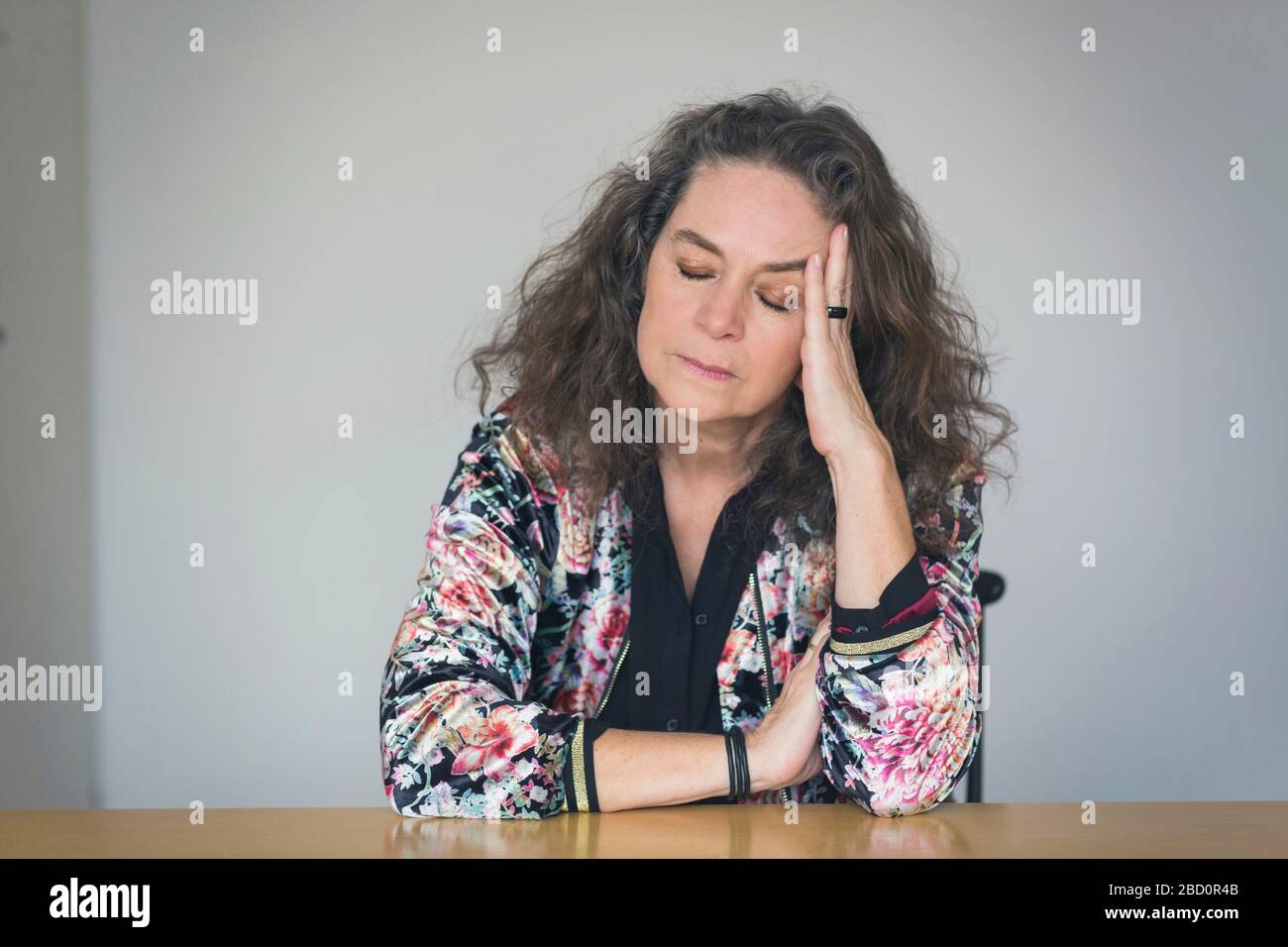 Depressed or unwell pretty middle-aged woman seated at a table indoors resting her head on her hand with closed eyes and a serious withdrawn expressio Stock Photo